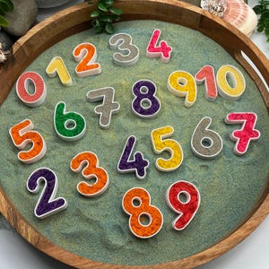 May include: A collection of colourful number-shaped containers filled with dyed rice, arranged on a circular wooden tray with green sand. The set includes digits zero through nine in various colours like red, orange, yellow, green, purple, and grey.
