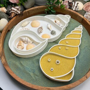 May include: A white seashell-shaped tray with compartments containing various seashells. Beside it is a seashell-shaped tray filled with yellow sand. The trays are on a green surface, inside a wooden tray, with more seashells around the edges.