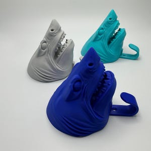 May include: Three shark head wall hooks in grey, teal, and blue. Each shark head features an open mouth with visible teeth and a curved hook extending from the back. The shark heads are moulded with detailed facial features and gills on the sides.
