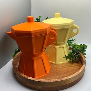 May include: Two geometric moka pots, one bright orange and one pale yellow, featuring a faceted octagonal body, a small lid knob, and a side handle. The pots are positioned together, showcasing a matte finish and a distinct, modern structural design.