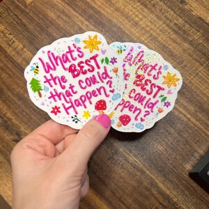 May include: Two die-cut holographic stickers featuring the pink text What's the BEST that could Happen? surrounded by colourful illustrations of a yellow sun, a red mushroom, a green tree, a bee, flowers, and blue clouds on a white background.