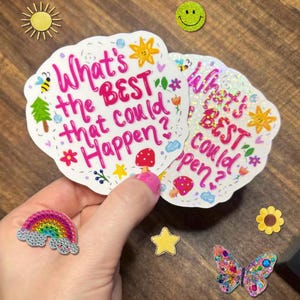 May include: Two die-cut stickers featuring the pink text What's the BEST that could happen? surrounded by small illustrations of a sun, bee, tree, mushroom, and flowers. One sticker has a holographic finish, while the other has a matte white finish.