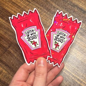 May include: Two die-cut stickers shaped like red condiment packets. Each features a white label with the text CATCH UP WITH JESUS and a red cross symbol. One sticker has a smooth finish, while the other has a holographic, sparkling effect.