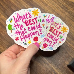 May include: Two die-cut stickers with a scalloped edge featuring the pink text What's the BEST that could happen? surrounded by colourful illustrations of a yellow sun, a green tree, a red mushroom, a bee, and small flowers on a white background.