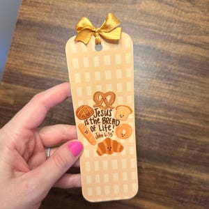 May include: A beige bookmark with a gold bow at the top. The bookmark features illustrations of baked goods and the text "Jesus is the Bread of Life! John 6:35". The background has a light brown checkered pattern.