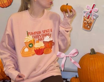 May include: A light pink crewneck sweatshirt featuring a graphic print of a pumpkin pie slice, a cupcake, a coffee drink, and a mug with the text PUMPKIN SPICE & EVERYTHING NICE. The sweatshirt is paired with dark brown trousers.