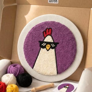 Op de afbeelding: Een punch needle kit met een cool kippenontwerp. De kit bevat een cirkelvormig paars en wit ontwerp, garen in verschillende kleuren, een punch needle tool en een schaar, allemaal gepresenteerd in een kartonnen doos.