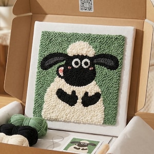 Op de afbeelding: Een knutselpakket met een schapenontwerp. De set bevat een canvas met een groene achtergrond en een schapenontwerp, garen in verschillende kleuren en een kartonnen doos. Het schaap heeft zwarte oren en een witte vacht.