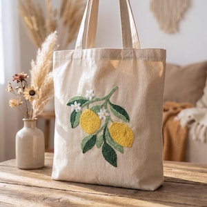 Op de afbeelding: Een beige canvas draagtas met een gedetailleerd geborduurd ontwerp van citroenen, bladeren en witte bloemen. De borduursels tonen heldergele citroenen en groene bladeren. De tas heeft twee lange handvatten en staat op een houten oppervlak.