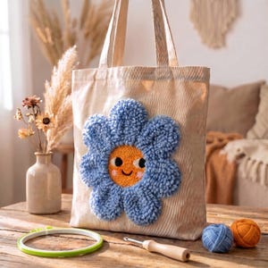 Op de afbeelding: Een beige draagtas met een groot, blauw, bloemvormig ontwerp met een oranje centrum en een lachend gezicht. De tas staat op een houten tafel met garenballen, een borduurring en een haaknaald.