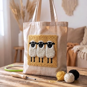 May include: A beige canvas tote bag featuring a square punch needle patch on the front. The patch displays three white sheep with black faces and legs against a textured mustard yellow background. The bag has two long shoulder straps.