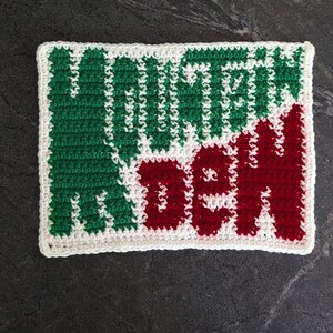 May include: A crocheted square with the words "Mountain Dew" in green and red. The text is centered on a white background with a white border. The square is likely a coaster or decorative piece.