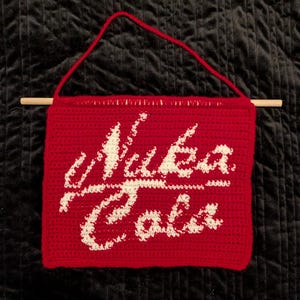 May include: A handmade, red crochet wall hanging with the words "Nuka Cola" in white. The sign is rectangular, with a wooden dowel at the top and a red yarn loop for hanging. The background is a dark, textured fabric.