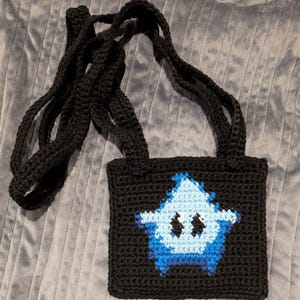 Puede incluir: Bolso negro de ganchillo con un diseño en forma de estrella azul y blanca en la parte delantera. El bolso tiene una correa larga para llevarlo al hombro. El bolso está hecho de un hilo grueso y tiene un aspecto texturizado.