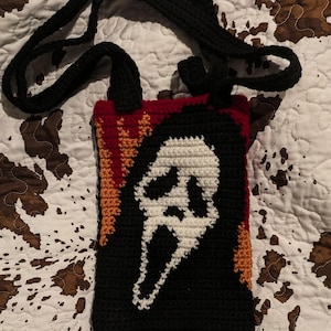 May include: A black, white, red, and orange crochet bag featuring the Ghostface mask from the Scream horror film series. The bag has a black strap and handles, perfect for carrying small items. The bag is handmade.
