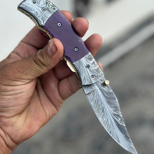 Puede incluir: Una navaja plegable con una hoja y un mango de acero estampado. El mango presenta una sección central morada vibrante y detalles metálicos decorativos. La hoja está parcialmente abierta, mostrando su intrincado diseño y filo afilado.