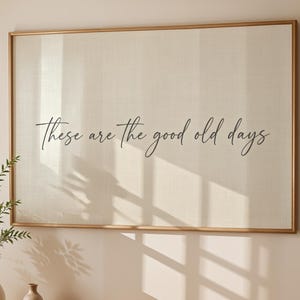 Peut inclure: Une impression sur toile encadrée avec le texte "These are the good old days" en écriture élégante. L'œuvre d'art est suspendue au-dessus d'une console en bois avec un vase de verdure, un petit vase et une tasse et une soucoupe. Une chaise en bois est à droite.