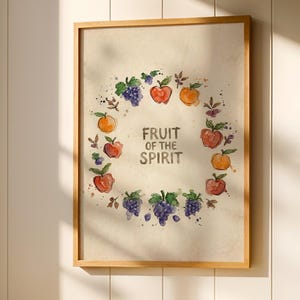 May include: A framed print with the text "FRUIT OF THE SPIRIT" surrounded by watercolor illustrations of fruit. A white marble table holds a white mug, a small ceramic cup, and a plate of oranges. The scene is set in a well-lit room.