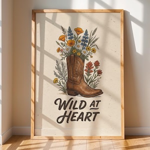 May include: A framed print featuring a brown cowboy boot filled with colorful wildflowers. The text "Wild at Heart" is written in a stylized font below the boot. The artwork is set against a cream-colored background.