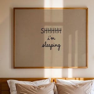 以下が含まれることがあります： SHHHHH i'm sleepingという黒い文字が中央に配置された長方形の額入りウォールアート。木製のベッドフレーム、白とベージュの枕、木製のナイトスタンド、質感のある茶色のランプがその下に配置されています。