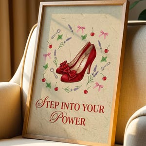 Puede incluir: Una impresión enmarcada con un par de zapatos de tacón rojos con lazos, rodeados de cerezas, lavanda y cucharas. El texto "Step Into Your Power" está escrito en una fuente decorativa. La obra de arte se exhibe en un sillón beige.