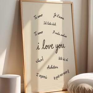 May include: A framed print with the words "I love you" in multiple languages. The print is in a light beige color with a wooden frame. The text is in black cursive. A small white cylindrical stool and a round beige cushion are in the foreground.
