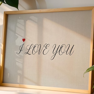 May include: A framed artwork with the words "I LOVE you" in elegant script, accompanied by a small red heart. The artwork is set against a neutral background and framed in light wood.