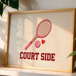 May include: A framed print featuring a pink tennis racket, ball, and heart illustration. The words "COURT SIDE" are printed in a bold, red font below the tennis-themed graphic. The artwork is set against a neutral background.