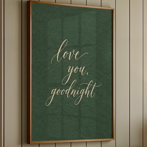 May include: A framed wall art piece with the words "love you, goodnight" in elegant cursive script on a dark green background. The artwork is displayed above a bed with a gray tufted headboard and white bedding. A lamp and a plant are on the nightstand.
