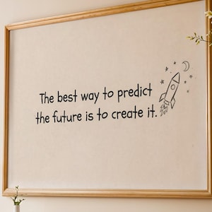 Op de afbeelding: Een ingelijst kunstwerk met de quote: "De beste manier om de toekomst te voorspellen is om hem te creëren." Het kunstwerk toont een raketillustratie. De lijst heeft een lichte houtkleur en de achtergrond is neutraal beige.