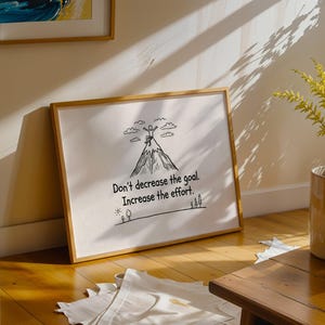 May include: A framed print with the text "Don't decrease the goal. Increase the effort." The artwork features a mountain with a person at the summit, and trees at the base. The frame is a light brown color.