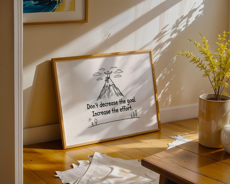 May include: A framed print with the text "Don't decrease the goal. Increase the effort." The artwork features a mountain with a person at the summit, and trees at the base. The frame is a light brown color.