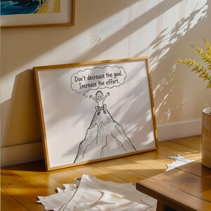 May include: A framed illustration with a motivational message. The artwork shows a person on a mountain peak with a thought bubble that reads, "Don't decrease the goal. Increase the effort." The frame is a light brown colour.