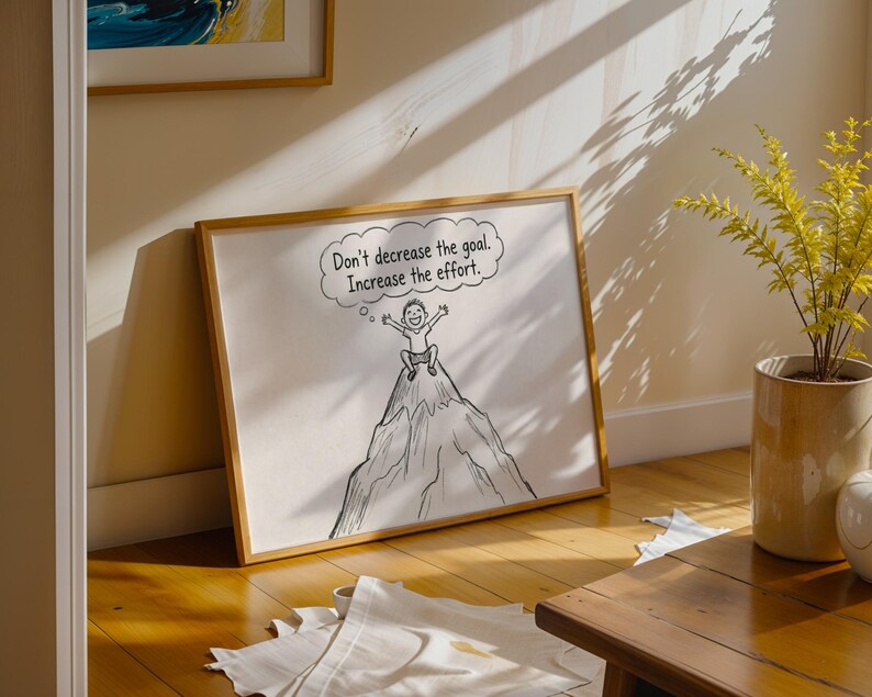 May include: A framed illustration with a motivational message. The artwork shows a person on a mountain peak with a thought bubble that reads, "Don't decrease the goal. Increase the effort." The frame is a light brown colour.