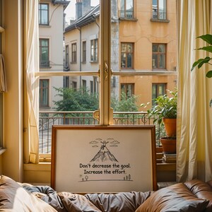 May include: A framed print with the text "Don't decrease the goal. Increase the effort." is displayed in front of a window. The print has a brown frame and a simple mountain illustration. The window overlooks a European cityscape.
