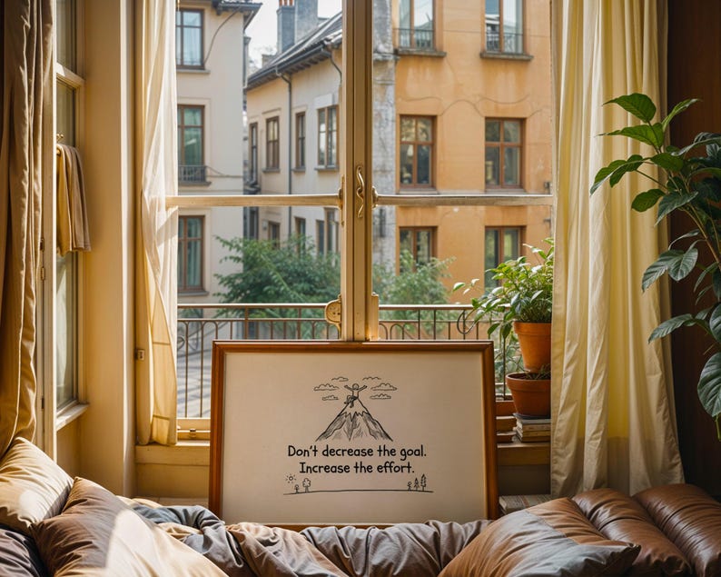 May include: A framed print with the text "Don't decrease the goal. Increase the effort." is displayed in front of a window. The print has a brown frame and a simple mountain illustration. The window overlooks a European cityscape.