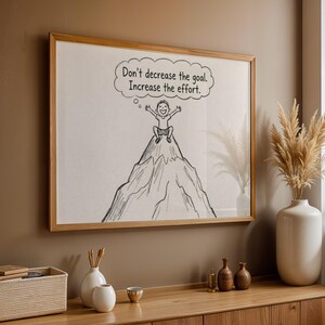 May include: A framed artwork with the text "Don't decrease the goal. Increase the effort." The illustration shows a person on a mountain peak. The frame is light brown wood. The artwork is displayed on a wall, with other decorative items.