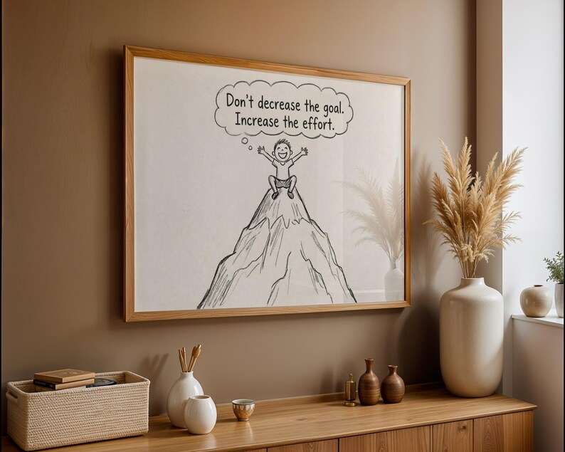 May include: A framed artwork with the text "Don't decrease the goal. Increase the effort." The illustration shows a person on a mountain peak. The frame is light brown wood. The artwork is displayed on a wall, with other decorative items.
