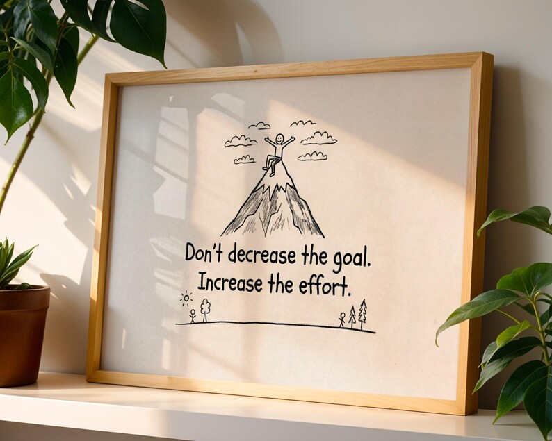 May include: A framed print with a motivational message. The artwork features a mountain with a person at the top, clouds, and the text "Don't decrease the goal. Increase the effort." The frame is made of light-colored wood.