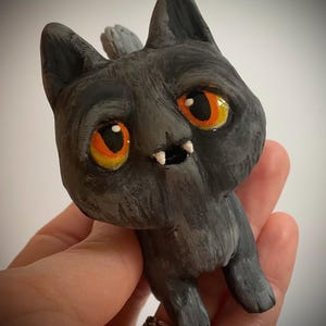 May include: A hand-painted, gray cat figurine with large orange and yellow eyes and small fangs. The cat has a small, gray tail and is posed in a standing position. The figurine is a unique, handcrafted item.