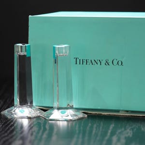 May include: Two clear glass candle holders with a faceted, hexagonal base and a cylindrical top. Each holder features a small blue sticker near the rim. Behind the holders sits a rectangular Tiffany & Co. box in a signature light blue color.