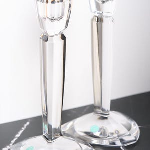 May include: Two clear crystal candlesticks with a faceted design. The candlesticks have a wide, geometric base and a slender stem leading to a cup-shaped top. The clear glass reflects light, creating a sparkling effect. Ideal for home decor.