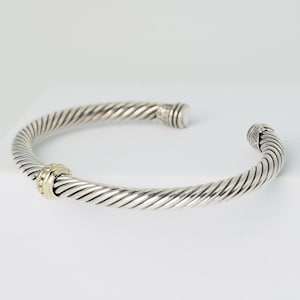 May include: A silver-toned cuff bracelet with a twisted rope design. The bracelet features a gold-colored accent with small, clear stones. The ends of the bracelet are open, and the overall design is elegant and minimalist.
