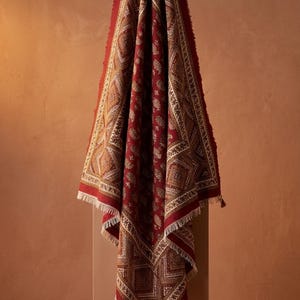 May include: A deep red textile featuring a complex, repeating geometric and paisley pattern in cream and brown tones. The fabric has a fringed edge and is draped vertically, showcasing a symmetrical border design and a central motif of small shapes.