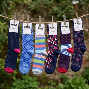 May include: Six pairs of crew socks hanging on a clothesline, each with a Koala Socks tag. Patterns include navy pinstripes, blue lattice, horizontal stripes in blue, green, and pink, red mushrooms on navy, red polka dots with blue stripes, and fireworks.