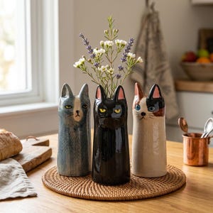 Peut inclure: Trois vases en céramique en forme de chat posés sur un tapis rond tressé. Le vase de gauche est bleu-gris, celui du centre est noir brillant et celui de droite présente un motif calicot crème, marron et noir. Le vase central contient des fleurs.