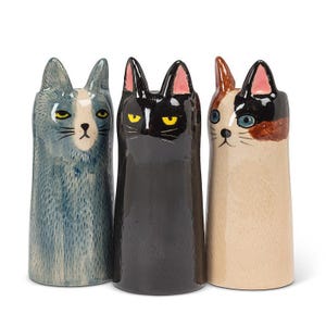 May include: Three ceramic cat-shaped vases standing side by side. The left vase is blue-grey with a white face patch. The centre vase is solid black with pink inner ears. The right vase is cream-coloured with brown and black patches on its head.