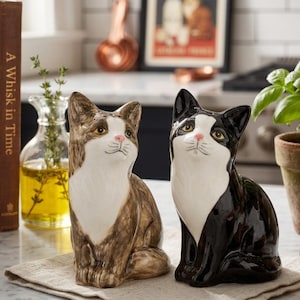 May include: Two ceramic cat figurines sitting upright. One cat features a brown and white tabby pattern, while the other is black and white. Both cats have white chests, pink noses, and yellow eyes, with a glossy finish and detailed facial features.