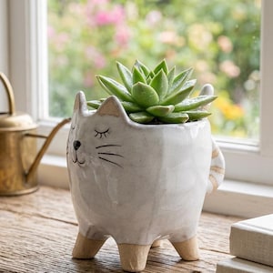 May include: A white ceramic cat-shaped planter with four tan feet, a painted black face with closed eyes and whiskers, and a striped tail. The planter holds a green succulent plant with pointed leaves, featuring a glossy finish on the body.
