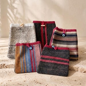 Peut inclure: Cinq pochettes rectangulaires en tissu avec fermetures à cordon. L'une est en tissu gris clair texturé, tandis que les autres présentent des motifs tissés en noir, rouge, bleu et beige. Chaque pochette est ornée d'une perle œil bleu ou d'un bouton en bois.